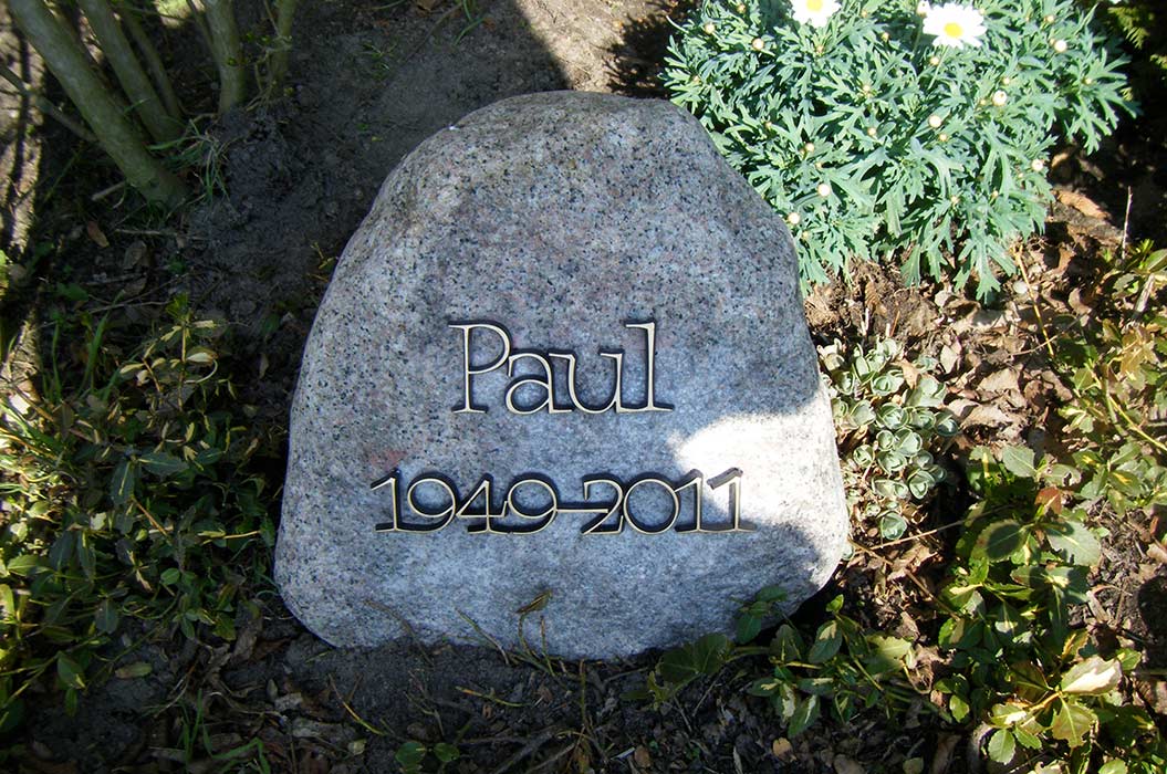 Ein Grabstein mit der Inschrift "Paul 1949–2011" inmitten von Pflanzen und Blumen.