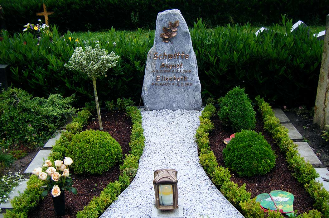 Ein gepflegtes Grab mit einem grauen Stein, weißen Blumen, einem Weg und einer Laterne.