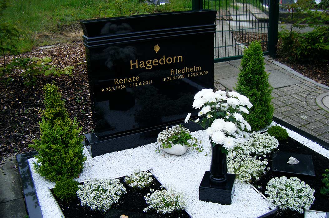 Schwarzer Grabstein mit Namen Hagedorn, weiße Blumen, gepflegtes Beet, grüner Friedhofshintergrund.