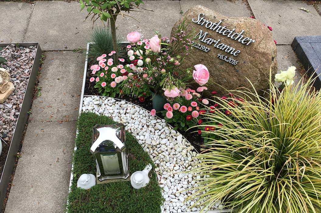 Grabstein mit Blumen, Laterne und Kerze auf gepflegtem Grab mit weißem Kies; Name nicht erkennbar.