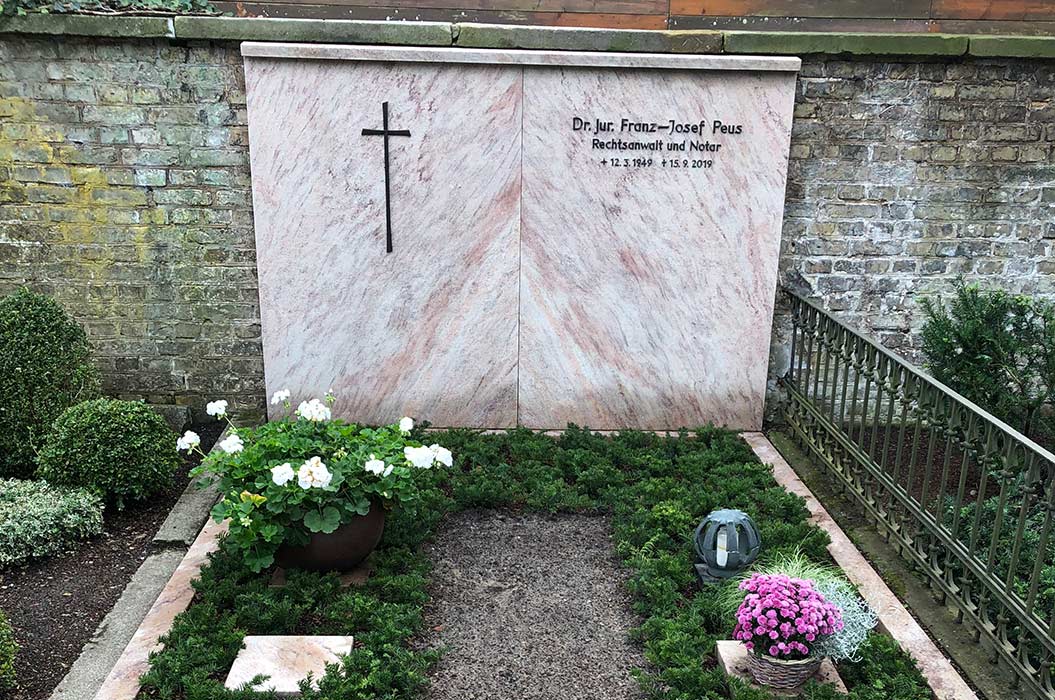 Grabstein mit Kreuz, Name Dr. jur. Franz-Josef Peus, umgeben von Blumen und grünem Laub.