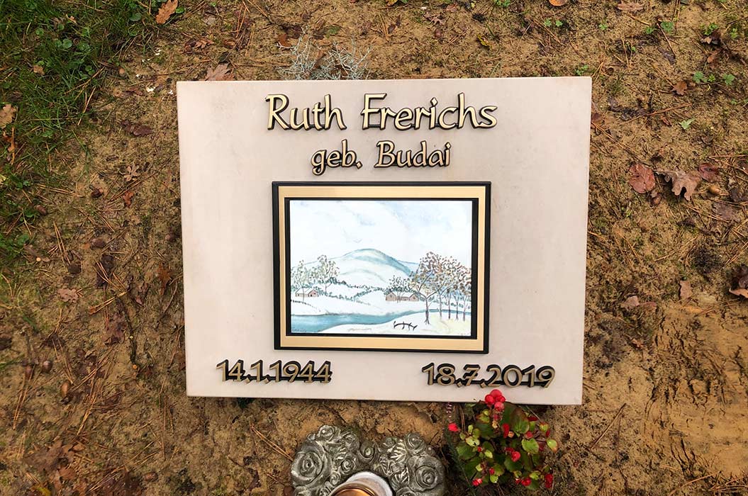 Grabstein mit Landschaftsbild, umgeben von braunem Boden. Kontrastiert von einer roten Blume.