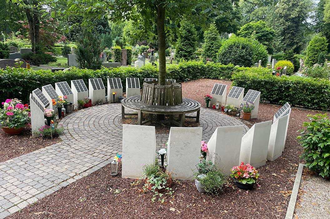 Runder Gedenkort mit Gedenkplatten, Blumen und Bänken um einen Baum in einem grünen Friedhof.