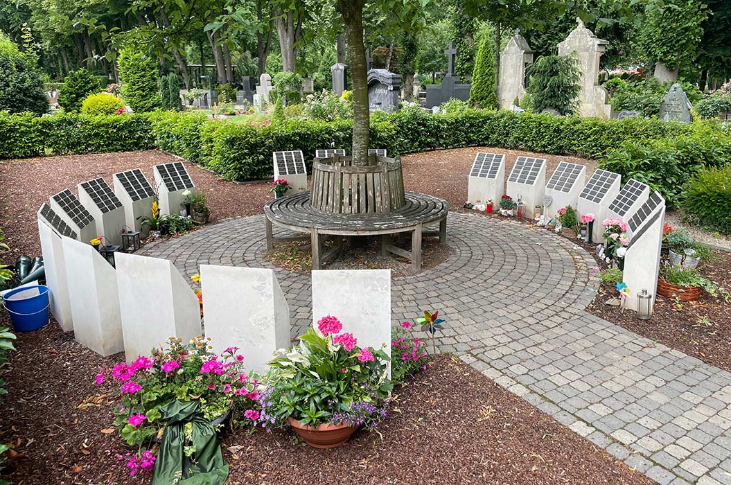 Kreisförmiger Friedhof mit Blumenbeeten und weißgrauen Grabsteinen um eine Bank unter Bäumen.