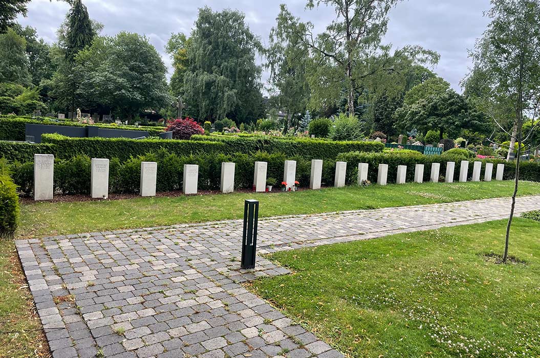 Reihe von Kriegsgräbern auf einem Friedhof mit Rasen und Bäumen, bewölkter Himmel.