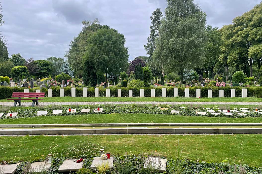 Ein gepflegter Friedhof mit Grabsteinen, Bäumen und einer roten Bank im grünen, bewölkten Park.