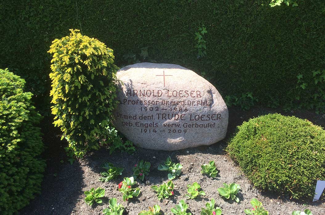 Ein Grabstein mit eingravierten Namen und Daten, umgeben von Pflanzen und einem kleinen Busch.