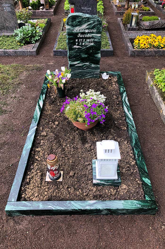 Grab mit schwarzem Grabstein, bunten Blumen, Laterne und Kerze in einer gepflegten Umgebung.