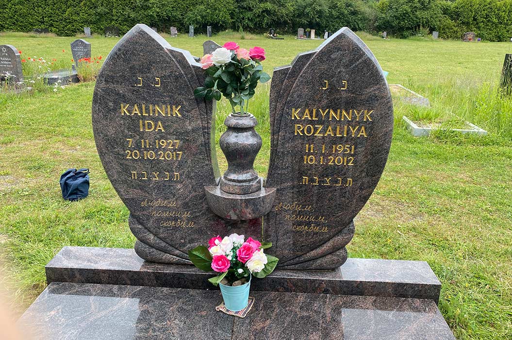 Gedenkstein mit Rosenstrauß, Inschrift auf Russisch und Hebräisch, auf grünem Friedhofshintergrund.