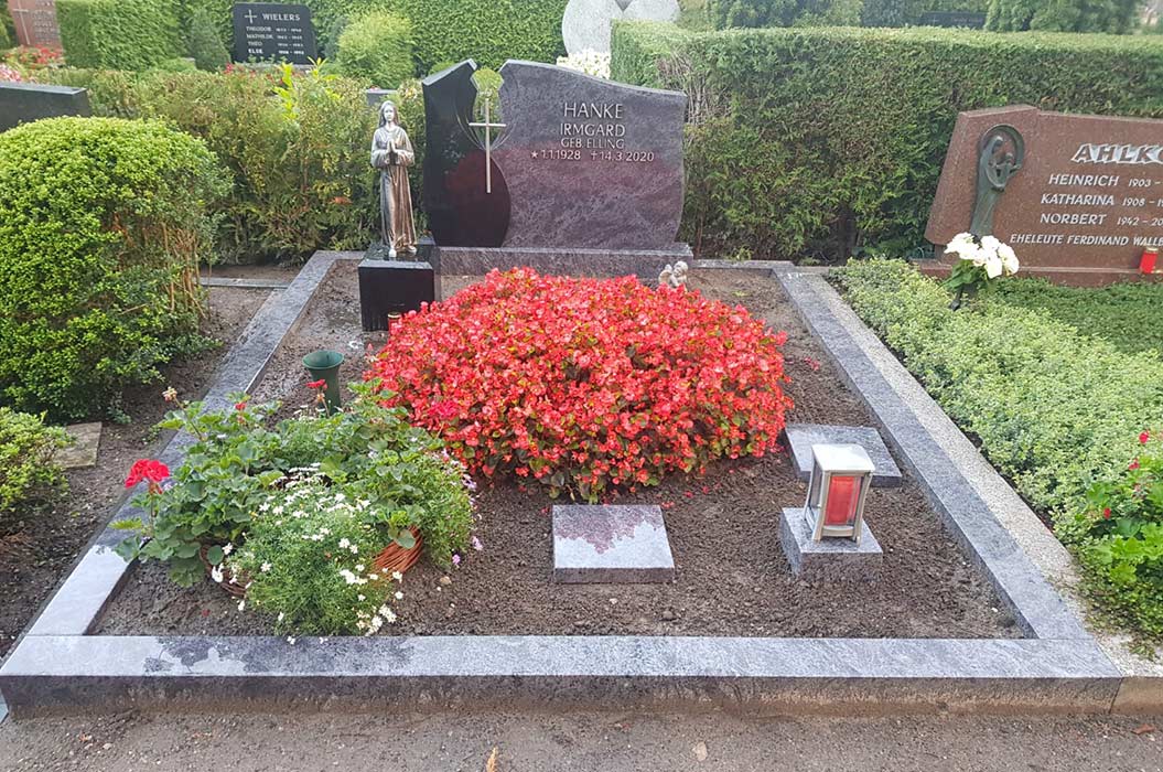 Grab mit rotem Blumenschmuck, Statue und Laterne auf einem gepflegten Friedhofsumfeld.