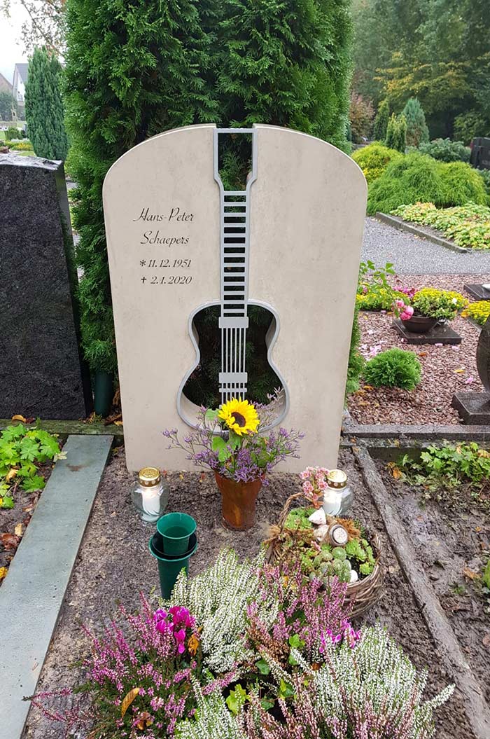 Grabstein in Gitarrenform mit Inschrift, umgeben von Blumen und Kerzen auf einem Friedhof.